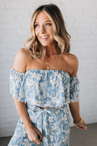 Blonde woman wearing a blue and white floral off the shoulder crop top with flowy overlay.