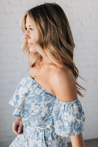 Blonde woman wearing a blue and white floral off the shoulder crop top with flowy overlay.