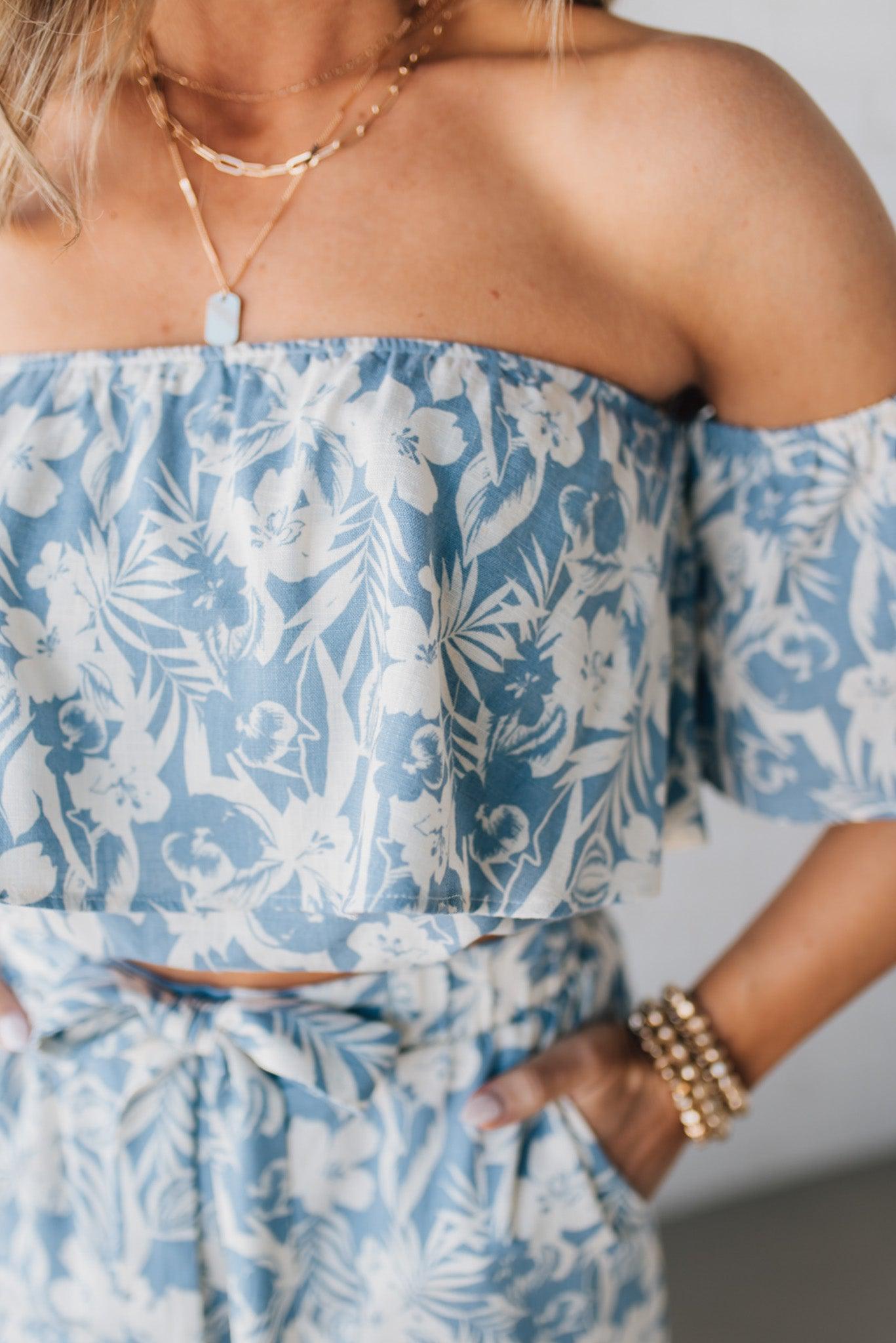 Blonde woman wearing a blue and white floral off the shoulder crop top with flowy overlay.