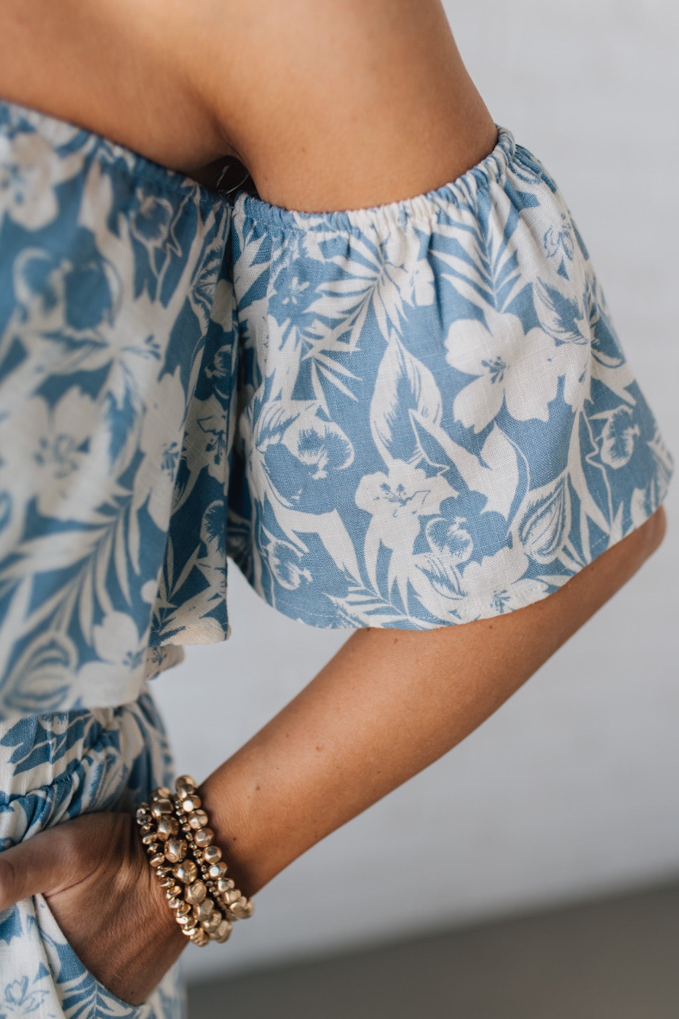 Blonde woman wearing a blue and white floral off the shoulder crop top with flowy overlay.