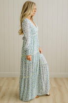 Woman wearing a long floral dress standing in a room with wooden flooring and white paneled walls.
