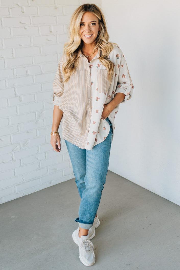 Woman wearing a floral blouse, jeans, and sneakers against a white brick wall.