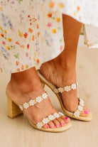 Beige sandals with floral embellishments worn with a floral dress on a neutral background
