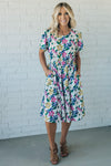 Women wearing a short sleeve midi dress that is Ivory with purple, pink and yellow flowers throughout.