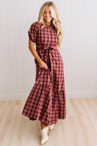 A woman modeling a red and dark brown plaid midi dress with puff sleeves, a tie waist, and a tiered ruffle hem, paired with white cowboy boots against a white paneled wall and light wood floor.