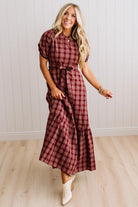A woman modeling a short-sleeve plaid midi dress in burgundy and brown, paired with cream ankle boots, standing in a room with light wood floors and white paneled walls.