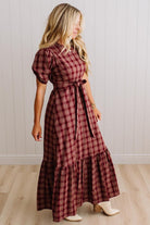 A woman modeling a long-sleeved, tiered maxi dress in a red and black plaid pattern, standing in a room with light wood floors and white paneled walls.