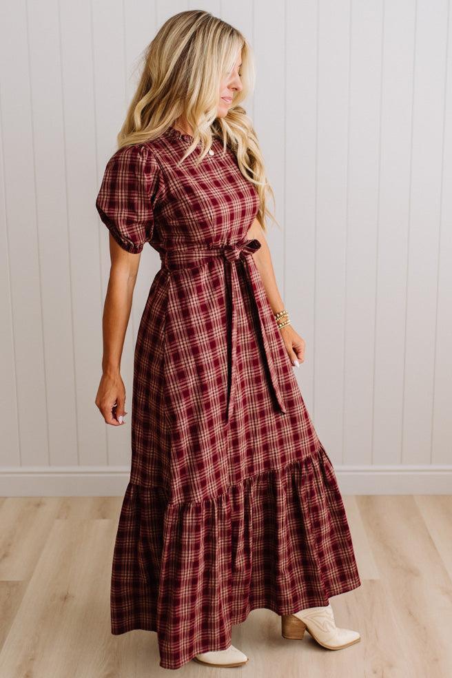 A woman modeling a long-sleeved, tiered maxi dress in a red and black plaid pattern, standing in a room with light wood floors and white paneled walls.
