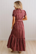 A woman wearing a red and dark plaid maxi dress, shown from the back in a room with white paneled walls and light wood floors.