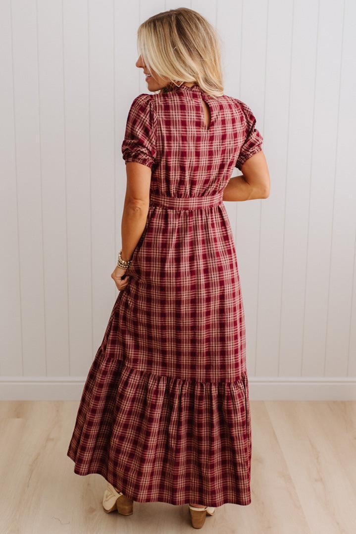 A woman wearing a red and dark plaid maxi dress, shown from the back in a room with white paneled walls and light wood floors.