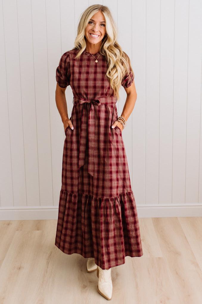 A woman modeling a short-sleeve plaid maxi dress in burgundy and brown tones, standing in front of a white paneled wall and light wood floor.