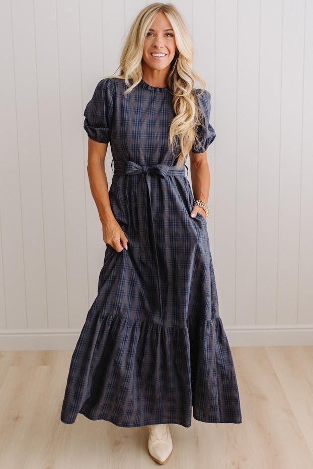 A woman modeling a navy blue plaid maxi dress with puff sleeves, a tie waist, and side pockets, standing in front of a white paneled wall and light wood floor.