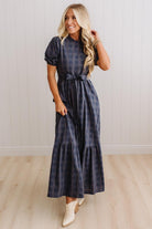A woman modeling a navy blue and gray plaid maxi dress with puff sleeves and a tie waist, paired with white cowboy boots, standing in front of a white paneled wall and light wood floor.
