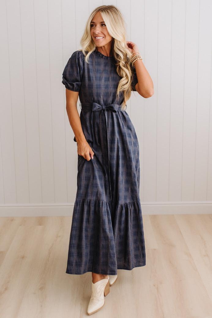 A woman modeling a navy blue and gray plaid maxi dress with puff sleeves and a tie waist, paired with white cowboy boots, standing in front of a white paneled wall and light wood floor.