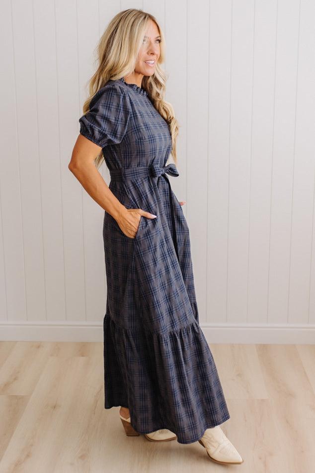 A woman modeling a navy blue and gray plaid midi dress with puff sleeves, side pockets, and a tie waist, standing in a room with light wood floors and white paneled walls.
