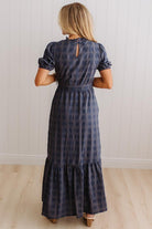 A woman viewed from behind wearing a navy blue plaid maxi dress with puff sleeves and a tiered skirt, standing in a room with white paneled walls and light wood floors.