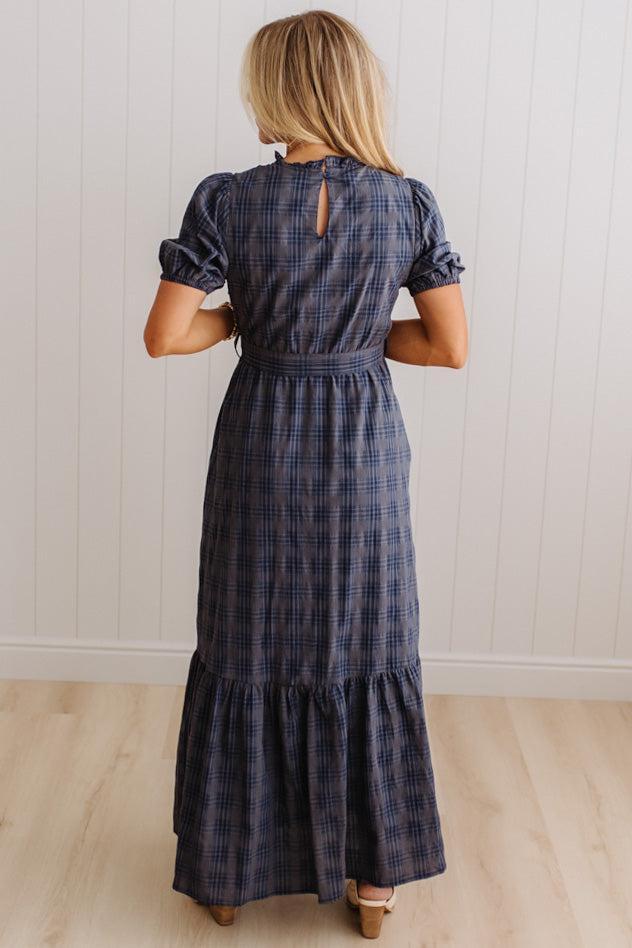 A woman viewed from behind wearing a navy blue plaid maxi dress with puff sleeves and a tiered skirt, standing in a room with white paneled walls and light wood floors.