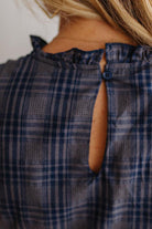 Close-up back view of a person wearing a navy and gray plaid top with a ruffled neckline and a small button closure at the back.