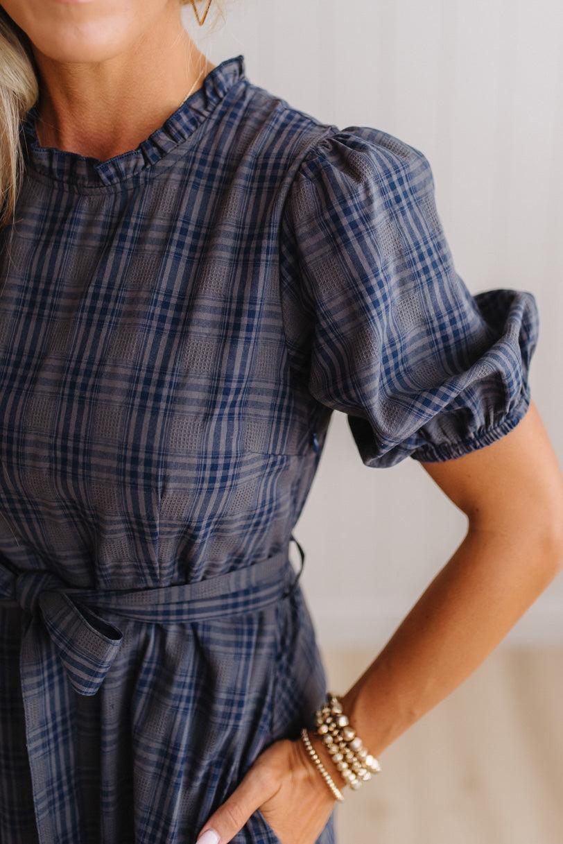 A close-up of a woman wearing a navy blue and gray plaid dress with puff sleeves and a matching waist tie, accessorized with gold bracelets.