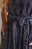 Close-up of a person wearing a blue and gray plaid dress with a matching tie belt, showing the waist and hands in pockets.