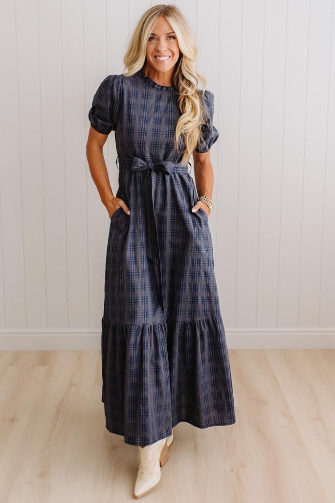 A woman modeling a navy blue and gray plaid maxi dress with puff sleeves, a tie waist, and side pockets, standing in front of a white paneled wall and light wood floor.