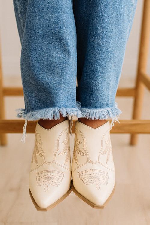 A close-up of a person's feet wearing cream-colored western-style pointed-toe ankle boots with tan embroidery, paired with frayed blue jeans.