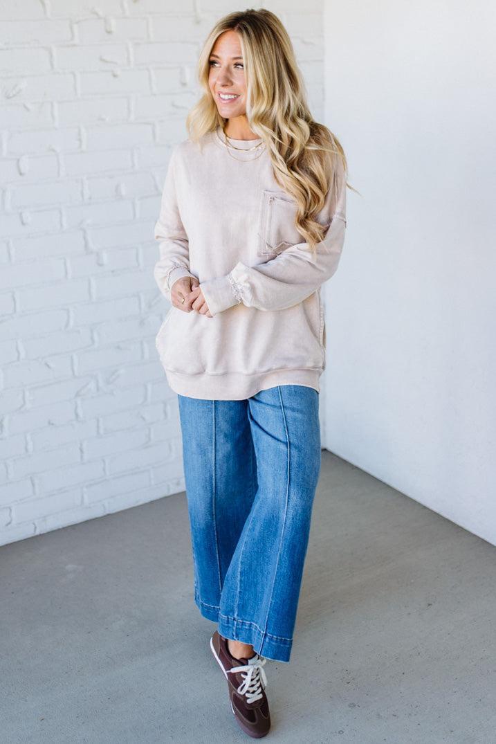 Woman wearing a beige sweatshirt and blue jeans against a white brick wall.