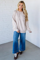 Woman wearing a beige sweatshirt and blue jeans standing against a white brick wall.
