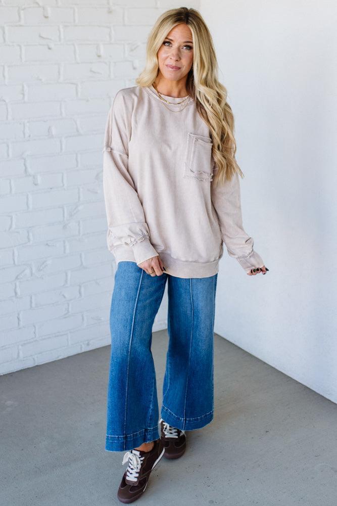 Woman wearing a beige sweatshirt and blue jeans standing against a white brick wall.