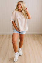 Woman wearing a beige oversized t-shirt and denim shorts standing on a wooden floor.