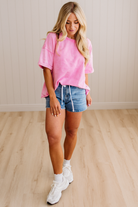 Woman wearing a pink oversized t-shirt, denim shorts, and white sneakers on a wooden floor.