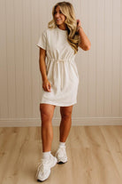 Woman wearing a white dress with white sneakers against a beige wall.