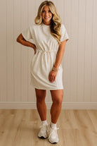 Woman wearing a white dress with a belt, standing against a beige paneled wall.