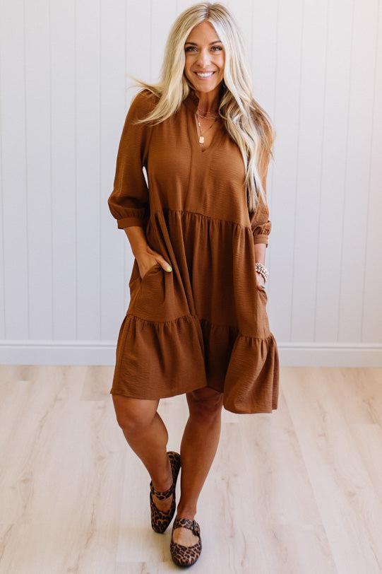 Blonde woman wearing a knee length solid dress with tiered skirt, v-neckline and side pockets in camel.