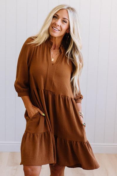 Blonde woman wearing a knee length solid dress with tiered skirt, v-neckline and side pockets in camel.