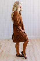 Blonde woman wearing a knee length solid dress with tiered skirt, v-neckline and side pockets in camel.