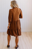 Blonde woman wearing a knee length solid dress with tiered skirt, v-neckline and side pockets in camel.