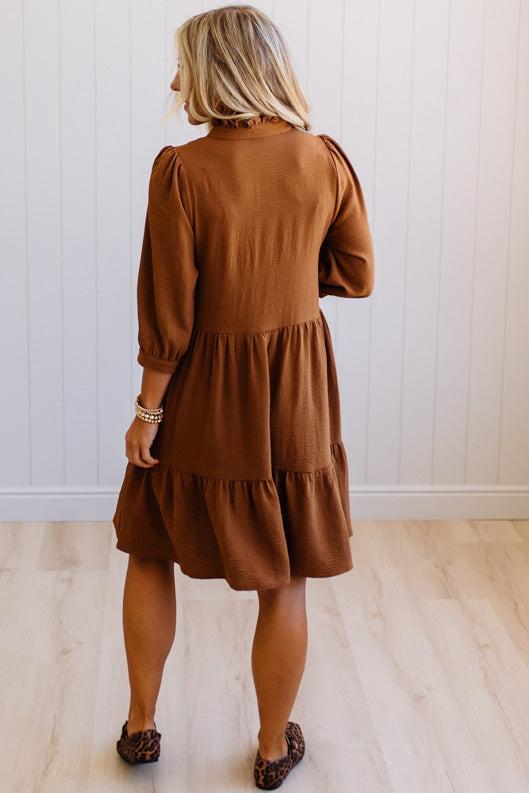 Blonde woman wearing a knee length solid dress with tiered skirt, v-neckline and side pockets in camel.