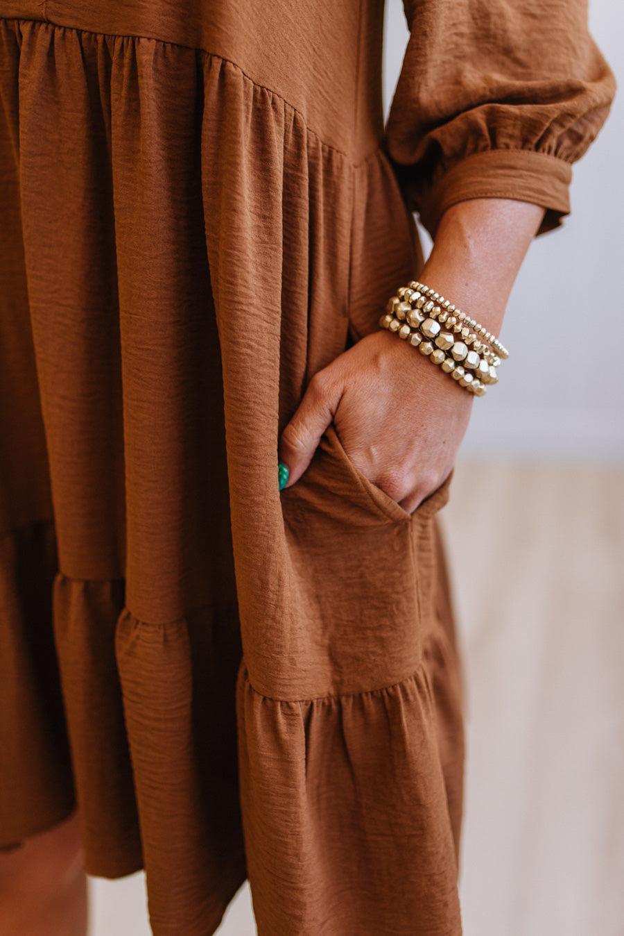 Blonde woman wearing a knee length solid dress with tiered skirt, v-neckline and side pockets in camel.