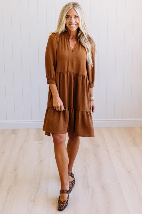 Blonde woman wearing a knee length solid dress with tiered skirt, v-neckline and side pockets in camel.