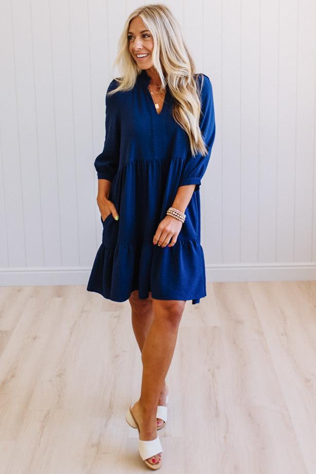 Blonde woman wearing a knee length solid dress with tiered skirt, v-neckline and side pockets in navy.