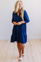 Blonde woman wearing a knee length solid dress with tiered skirt, v-neckline and side pockets in navy.