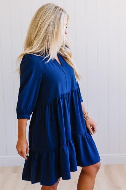 Blonde woman wearing a knee length solid dress with tiered skirt, v-neckline and side pockets in navy.