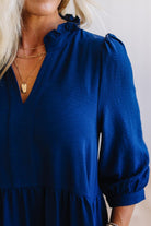 Blonde woman wearing a knee length solid dress with tiered skirt, v-neckline and side pockets in navy.