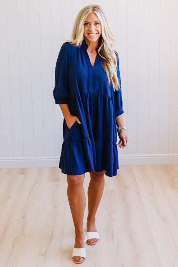 Blonde woman wearing a knee length solid dress with tiered skirt, v-neckline and side pockets in navy.