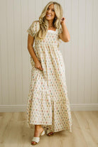 Woman wearing a floral dress standing against a light wooden paneled wall.