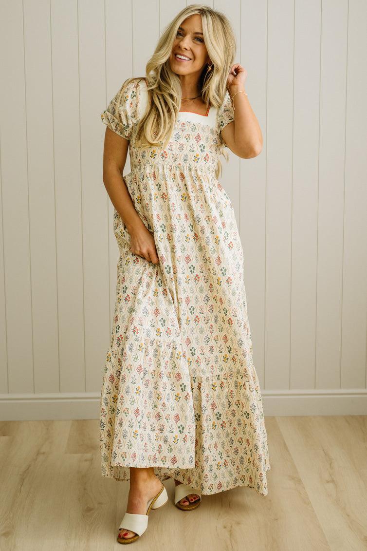 Woman wearing a floral dress standing against a light wooden paneled wall.