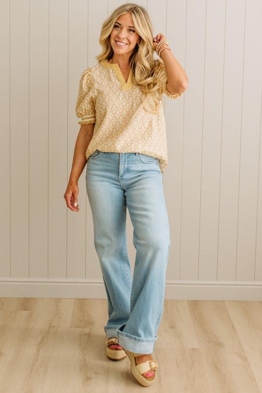 Woman wearing a beige patterned top and light blue jeans against a white paneled wall.