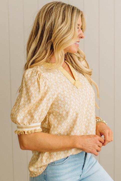 Woman wearing a beige patterned blouse and light blue jeans against a neutral background