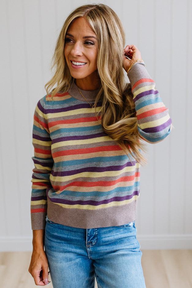 Blonde Woman Modeling a Wool Blended Multi Color Stripe Sweater Standing on a Light Wood Floor with Cream Backdrop.